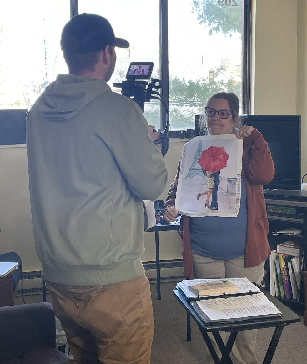 Young woman named Maria holding art work. A man is using a camera to film a video of her.